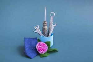 Creative vase with tools screwdriver wrench. On a colored background a pink rose a blue tie and a vase with tools. Minimalism for Father's Day. Close up, monochrome.Still life photo