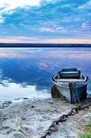 An old wooden boat is moored to the bank with a rusted iron chain on the bank of a calm river against the morning horizon. photo