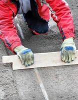 el trabajador, arrodillado, nivela manualmente la base de arena con una tabla de madera para el futuro revestimiento de aceras. imagen vertical. foto