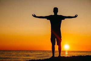 Silueta de un hombre parado y contemplando en la playa en el fondo del mar y el atardecer arrojando sus manos hacia un lado foto