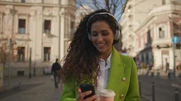 Bastante joven escuchando música con el teléfono inteligente en la calle y sosteniendo café para llevar video