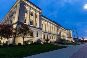 Kenosha, Wisconsin, County Courthouse at night. Brilliant blue sky.  Courthouse lights shining on the architecture.  Wide sidewalk lined with bricks and street lights. Tree branches glowing in light photo