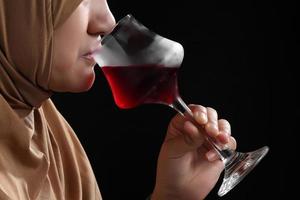 Close up muslim woman in a hijab drink red wine on black background photo
