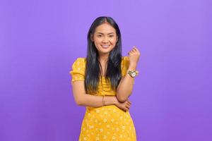 Smiling young Asian woman wearing yellow dress and looking confident on purple background photo