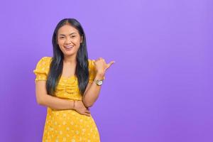 Portrait of smiling young Asian woman pointing thumb at copy space on purple background photo