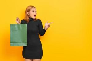 Surprised young Asian woman holding shopping bags and pointing finger at copy space isolated over yellow background photo