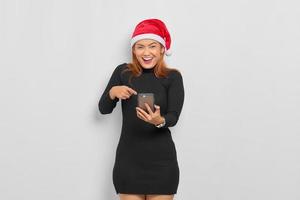 Cheerful young Asian woman in Santa Claus hat pointing fingers at mobile phone isolated over white background photo