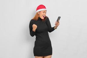 Excited young Asian woman in Santa Claus hat looking at mobile phone and celebrate luck isolated over white background photo