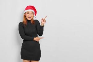 Smiling young Asian woman in Santa Claus hat pointing finger at copy space isolated over white background photo