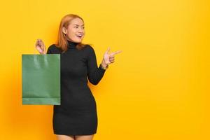 Cheerful young Asian woman holding shopping bags and pointing finger at copy space isolated over yellow background photo