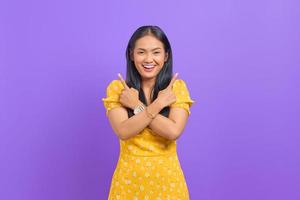 Sonriente joven mujer asiática apuntando hacia un lado y mirando a la cámara sobre fondo púrpura foto