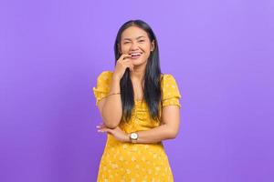 Portrait of smiling young Asian woman standing and biting her nails on purple background