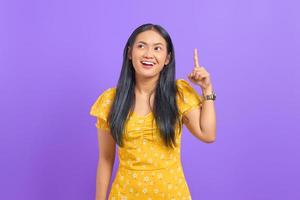 Smiling young Asian woman pointing fingers up and having an idea on purple background photo