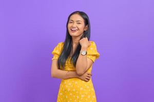 Smiling young Asian woman wearing yellow dress and looking confident on purple background photo