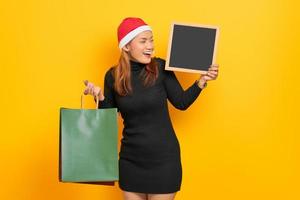 Sonriente joven mujer asiática en santa claus hat sosteniendo bolsas de compras y pizarra aislado sobre fondo amarillo foto