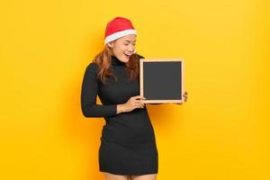 Portrait of smiling young Asian woman in Santa Claus hat holding chalkboard isolated over yellow background photo