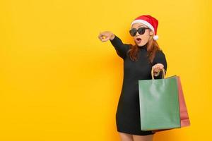 Surprised young Asian woman in Santa Claus hat and sunglasses holding shopping bags, pointing finger at copy space isolated over yellow background photo