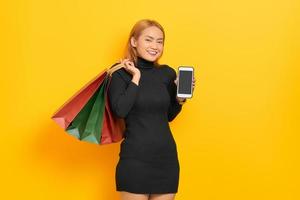 Smiling young Asian woman showing blank screen mobile phone and holding shopping bags isolated over yellow background photo