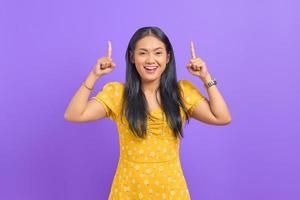 Sonriente joven mujer asiática apuntando con el dedo hacia arriba en el espacio de la copia sobre fondo púrpura