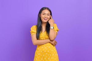 Smiling young Asian woman wearing yellow dress and looking confident on purple background photo