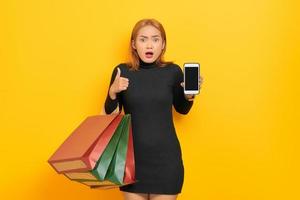 Surprised young Asian woman holding blank screen mobile phone and gesturing thumbs up, holding shopping bags isolated over yellow background photo