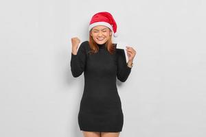 Cheerful young Asian woman in Santa Claus hat showing a blank card and celebrate luck isolated over white background photo