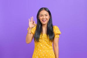 Alegre joven asiática hace un gesto bien, demuestra el símbolo de aprobación sobre fondo púrpura foto