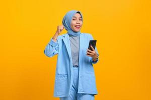 Cheerful young Asian woman holding mobile phone and celebrating success over yellow background photo