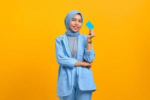 Smiling young Asian woman holding credit card in hand and looking at camera over yellow background photo