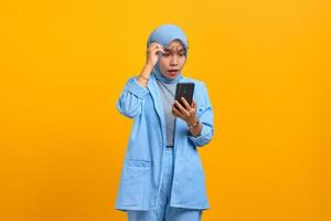 Shocked young Asian woman using mobile phone and taking of glasses isolated over yellow background photo