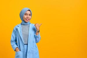 Cheerful young Asian woman pointing fingers aside at copy space isolated over yellow background photo