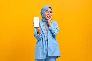 Cheerful young Asian woman showing smartphone blank screen with hand on cheek over yellow background photo