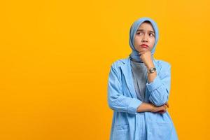 Pensive young Asian woman thinking about question with hands on chin over yellow background photo