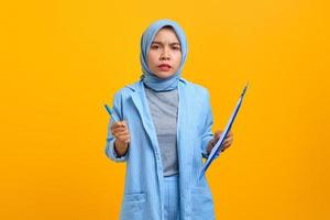 Angry young Asian woman holding pen and document folder over yellow background photo