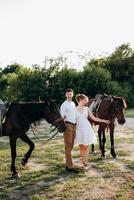 girl in a white sundress and a guy in a white shirt on a walk with brown horses photo