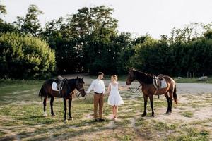 girl in a white sundress and a guy in a white shirt on a walk with brown horses photo