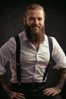 portrait of a brutal man with a beard and long hair he looks at the camera and smiles dressed in a shirt and jeans with suspenders photo