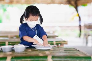 Linda estudiante con máscara en forma de 4d está estudiando en el aula en una granja natural. La niña usa sus dos manitas para mezclar polvo de tiza blanca en una placa de zinc sobre una mesa de bambú. Aprendiendo con nueva normalidad. foto