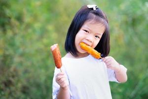 Child holds two sausages in her hands. Cute girl was eating delicious skewer meal. A 4-5 year old kid wears white shirt. Children is hungry. Green nature background. Empty space to enter text.