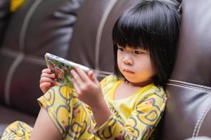 Child is watching video with smartphone on brown leather sofa. Apartment life. photo