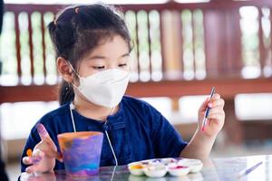 Child doing watercolor art. Kid holding brush. Children paint crafts on clay plant pots. Girl wear mask to prevent small toxic dust air pollution PM2.5 and spread virus. New Normal. photo