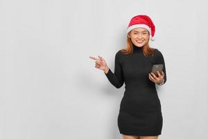 Sonriente joven mujer asiática con sombrero de santa claus sosteniendo teléfono móvil y señalando con el dedo en el espacio de copia aislado sobre fondo blanco. foto