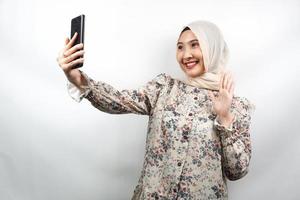 Hermosa joven musulmana asiática sonriendo confiada, entusiasta y alegre con las manos sosteniendo el teléfono inteligente, llamando a alguien, haciendo videollamadas, tomando selfie, aislado sobre fondo blanco. foto