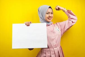 Bastante joven mujer musulmana alegre sosteniendo pancarta vacía en blanco, cartel, tablero blanco, tablero de letrero en blanco, tablero de anuncio blanco, presentando algo en el espacio de la copia, promoción foto