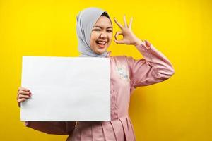 Bastante joven mujer musulmana alegre sosteniendo pancarta vacía en blanco, cartel, tablero blanco, tablero de letrero en blanco, tablero de anuncio blanco, presentando algo en el espacio de la copia, promoción foto