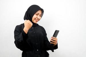 Hermosa joven musulmana asiática con las manos sosteniendo el teléfono inteligente, sonriendo confiado, entusiasta y alegre, con las manos apretadas, puñetazos, victoria, éxito, buen trabajo, aislado sobre fondo blanco.
