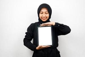 Beautiful young asian muslim woman smiling, hand holding tablet with white or blank screen, promoting app, promoting product, presenting something, excited and cheerful, isolated on white background photo