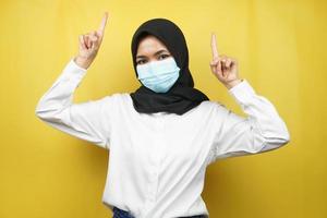 Mujer musulmana con máscara médica, mano apuntando al espacio vacío, mano apuntando hacia arriba presentando algo, aislado sobre fondo amarillo foto