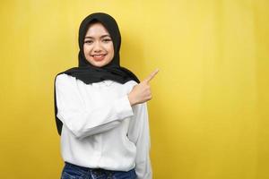 Beautiful young asian muslim woman with hands pointing empty space presenting something, smiling confident, enthusiastic, cheerful, looking at camera, isolated on yellow background photo