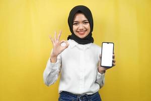 Beautiful young asian muslim woman smiling confidently and excitedly with hands holding smartphone, promoting application, ok sign hand, good job, success, isolated on yellow background